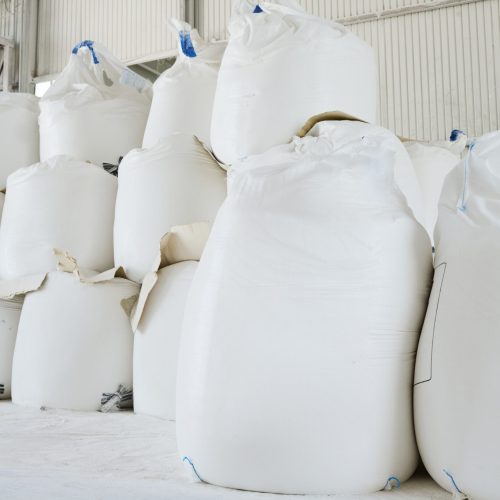 Heaps of huge white sacks containing loose raw materials prepared for production process in spacious warehouse or storage room